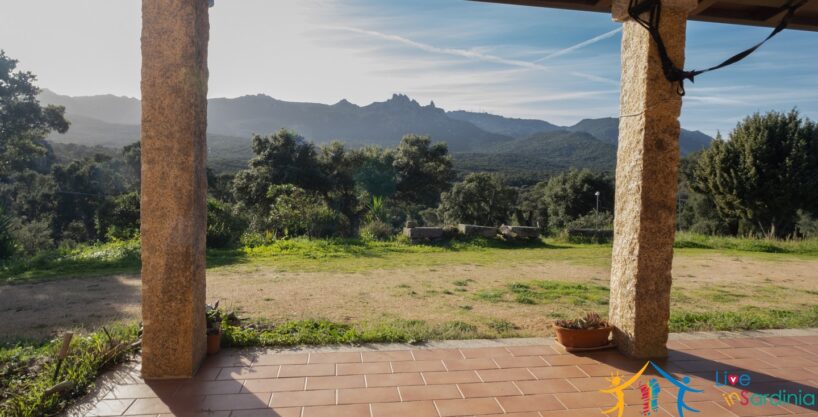 Covered veranda with terracotta tiles and granite columns, looking out over a wide, natural landscape and majestic mountain ridge.